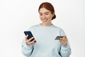 Image of smiling young redhead woman register her credit card in application, looking at mobile phone screen, holding Royalty Free Stock Photo