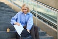 Image of smiling young woman, student packing her laptop in backpack, sitting on public stairs with cup of coffee, hurry