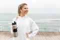 Image of smiling young woman using earpods and drinking water from bottle while walking near seaside Royalty Free Stock Photo