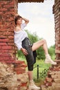 Image of a stylish beautiful woman in a white shirt and leather skirt in a park against the background of a destroyed building.