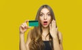 Image of surprised young lady standing over yellow background and holding debit card in hands. Looking at camera Royalty Free Stock Photo