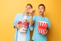Image of two excited beautiful teenagers, guys watching an interesting movie and eating popcorn on a yellow background