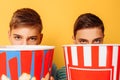 Image of two frightened teenagers, guys watching a horror movie and hiding behind a bucket of popcorn on a yellow background