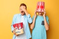 Image of two frightened teenagers, guys watching a horror movie and hiding behind a bucket of popcorn on a yellow background