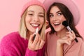 Image of two joyful women smiling and applying makeup with cosmetics