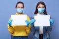 Image of two young women wearing medical disposable masks and medical gloves holding white blank papers in hands, copy space for Royalty Free Stock Photo