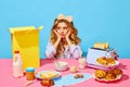 Image of upset girl in homewear and bow propping up cheeks and looking away from food on pink tablecloth over light blue Royalty Free Stock Photo