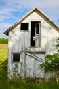 Vertical shot of abandoned farmhouse in grassy field near farmland with busted windows and door