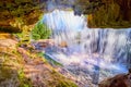 View behind and inside of waterfall cave of orange and lichen growth with sun hitting the water