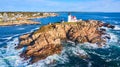 Waves crash over rocks aerial over Maine island with lighthouse and view of homes on mainland