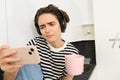 Image of woman with disappointed, reluctant face expression, watching boring show or movie on smartphone, wearing