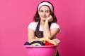 Image of woman stands near huge pink basin with fresh clothes, blankets, towels and other home textile, finishes washing, has