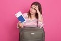 Image of woman in striped shirt over pink background, sitting near her suitcase, holding documents in hands, keeps hand on Royalty Free Stock Photo