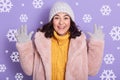 Image of woman wearing stylish eco fur coat, yellow sweater and cap, looking at camera and keeping hands up in studio with