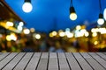 Image of wooden table in front of decorative outdoor string lights hanging on electricity post in night market with blur people.