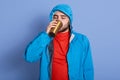 Image of young attractive bearded man poses isolated over blue background, drinks tea or coffee from thermo mug, wears jacket with Royalty Free Stock Photo