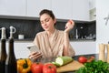 Image of young beautiful woman, holding tomato, sitting in kitchen with smartphone, chopping board and vegetables on Royalty Free Stock Photo