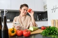 Image of young beautiful woman, holding tomato, sitting in kitchen with smartphone, chopping board and vegetables on Royalty Free Stock Photo