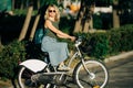 Image of young blonde in sunglasses and long denim skirt standing on bike next to green bushes in city Royalty Free Stock Photo