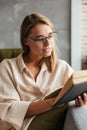 Image of young concentrated woman reading book while sitting on couch