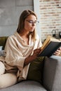 Image of young concentrated woman reading book while sitting on couch