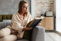 Image of young concentrated woman reading book while sitting on couch