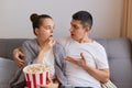 Image of young couple sitting on couch and watching movie, woman wrapped in blanket eating popcorn, husband looking at her with Royalty Free Stock Photo