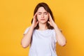 Image of young good looking brunette female stands with closed eyes isolated over yellow background, keeps hands on temples, has Royalty Free Stock Photo