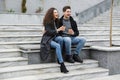 Image of young satisfied man and woman 20s in warm clothes, drinking takeaway coffee while sitting on bench outdoor