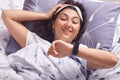 Image of young woman in bed lying on gray pillow under white blanket, wants to know what time is it, looks at her smart watch, Royalty Free Stock Photo