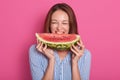 Image of young woman biting big slice of sweet watermelon while smiling and looking at camera, posing  over rose Royalty Free Stock Photo