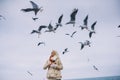 Image of young woman feeds seagulls on the sea. Pretty female wearing coat, scarf and watching flying seagulls by the sea in the Royalty Free Stock Photo