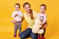 Imagine of young beautiful mother with little twin girls posing isolated over yellow background, children wants to play with their