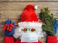 Imitation of a comic image of a frightened Santa Claus in a hat made of a medical mask, gloves, glasses on a wooden background.