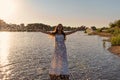 An immature young girl in a white wet dress is standing in a river and sprinkles water droplets up in the rays of the rising sun