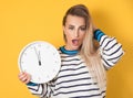 Impatient woman with angry face expression holding clock, isolated on yellow background. Stressed girl showing wall clock