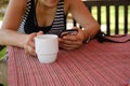 Impersonal woman using smartphone and drinking tea from the white cup at the table with the striped tablecloth.