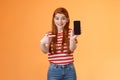 Impressed excited happy redhead woman smiling broadly, hold smartphone, pointing cellphone screen, thrilled amazed
