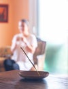 Incense stick burning with a woman sitting crosslegged in the background in a cozy room