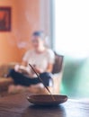 Incense stick burning with a woman sitting crosslegged in the background in a cozy room