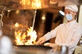 Skillful chef in face mask holding wok in flames