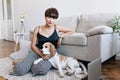 Incredible brunette lady wears elegant wristwatch posing beside sofa stroking cute beagle. Indoor portrait of blissful