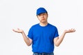 Indecisive clueless asian young delivery man in blue uniform, shrugging and looking away puzzled, spread hands sideways