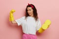 Independent strong housewife shows her muscle, wears yellow gloves and holds a sponge, isolated over pink wall