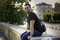 Indian arab or hispanic young man in black shirt with a shoulder strap bag and sunglasses sitting on a wall smiling while looking Royalty Free Stock Photo