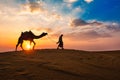 Indian cameleer camel driver with camel silhouettes in dunes on sunset. Jaisalmer, Rajasthan, India