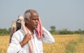 Indian farmer with hand hoe on his shoulder holding towel to his face due to high temperature while working on agriculture land