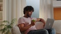 Indian freelancer man eating apple while workg witin laptop indoors. Young hungry guy sitting on comfortable couch