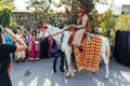 Indian groom ridding white horse with yellow and red pattern fabric, flower necklace and red turban with guests.