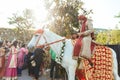 Indian groom ridding white horse with yellow and red pattern fabric, flower necklace and red turban with guests.